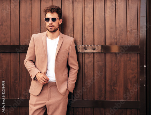 Handsome confident stylish hipster lambersexual model.Sexy modern man dressed in elegant beige suit. Fashion male posing in the street near wooden fence wall in city at sunset photo