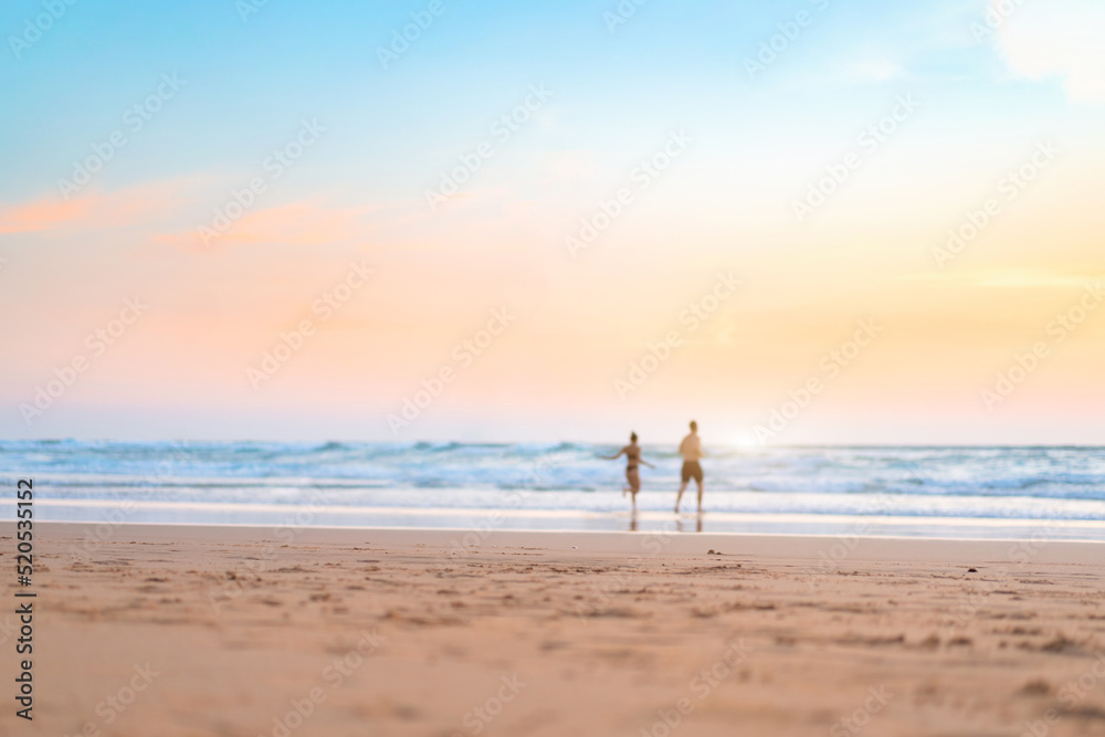 Couple running on the beach. Happy couple go to swim in ocean at sunset. Blurred summer vacation background. Defocused man and woman run on sandy sea beach. Summertime.