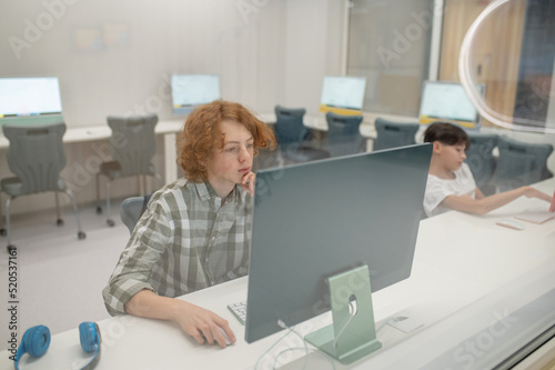 A ginger schoolboy sitting at the computer at school and looking involved