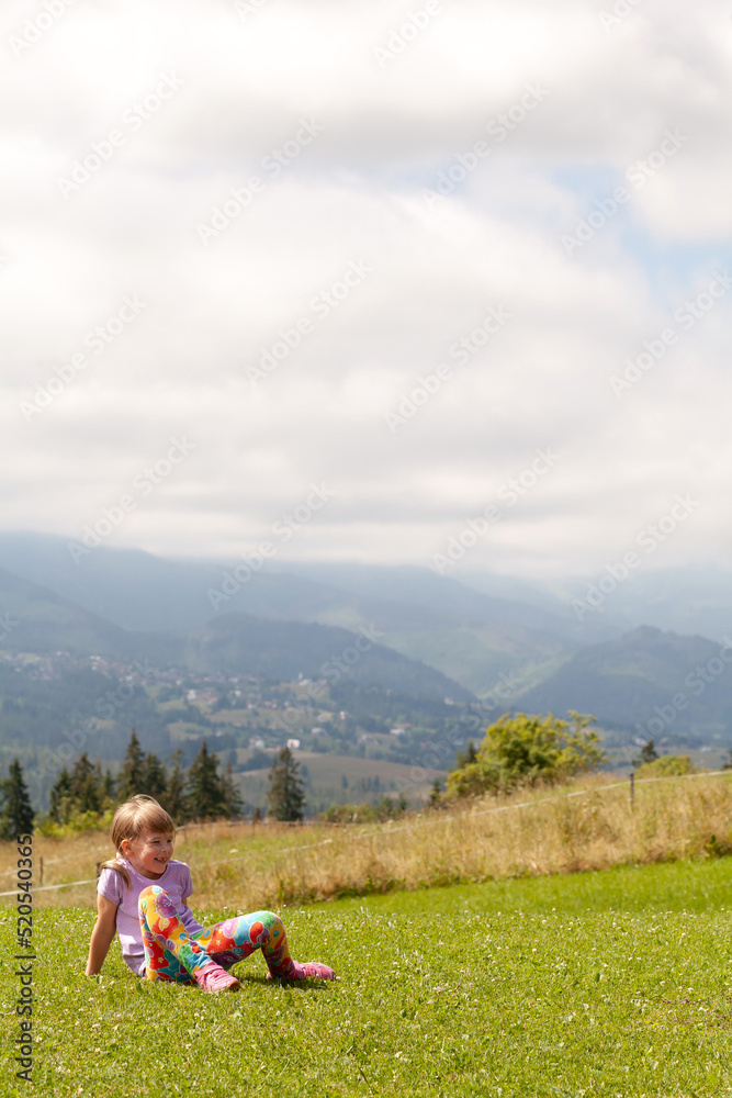 Happy cheerful elementary school age girl sitting on the ground on a green grass field alone laughing, outdoors shot. Copy space poster background, one person. Mountains, countryside, rural area