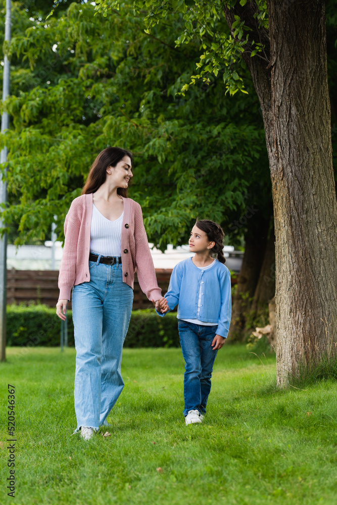 happy woman and girl holding hands and looking at each other while walking in park happy woman and girl holding hands and looking at each other while walking in park