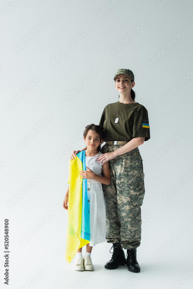 Full length of smiling woman in camouflage uniform hugging daughter with ukrainian flag on grey background Full length of smiling woman in camouflage uniform hugging daughter with ukrainian flag on grey background