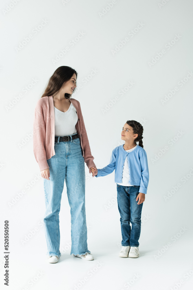 full length of woman and girl in jeans holding hands and smiling at each other on grey background full length of woman and girl in jeans holding hands and smiling at each other on grey background
