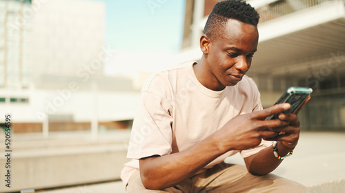 Young african student sits outside of university, uses cellphone, taps on smartphone screen
