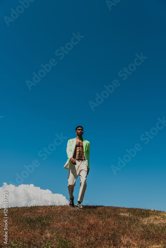 full length of african american man in trousers and blazer walking in field under blue sky
