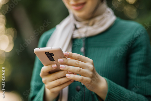 Cropped view focus on smartphone. Arabic muslim young women wearing hijab listening to music with headset, having coffee break outdoor on park background.