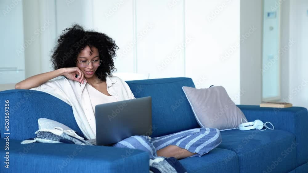Pretty curly haired woman in eyeglasses working on laptop while sitting on the sofa at home Pretty curly haired woman in eyeglasses working on laptop while sitting on the sofa at home
