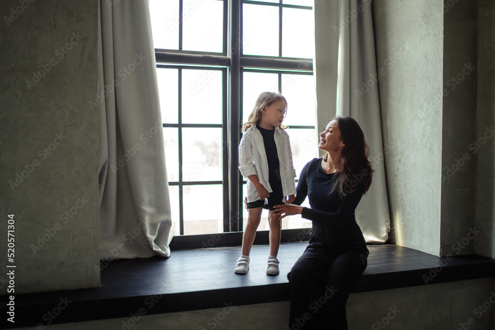 happy mother and daughter having fun and hugging at home In a dark room sitting on a large window happy mother and daughter having fun and hugging at home In a dark room sitting on a large window