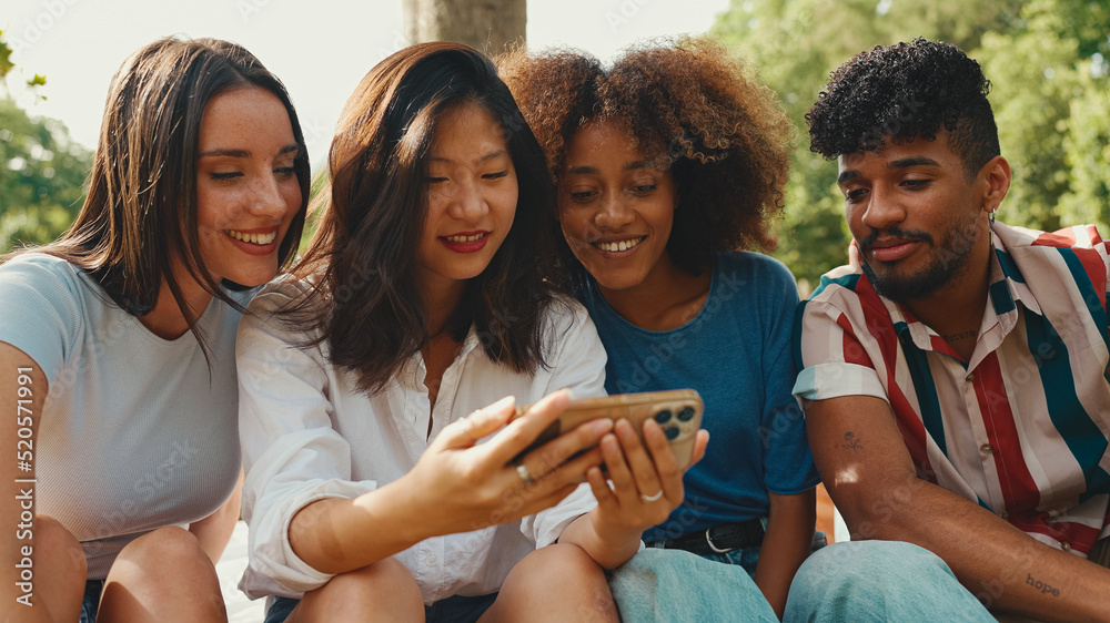Fototapeta premium Happy, smiling multiethnic young people at picnic on summer day outdoors. Group of friends talking, using cellphone while relaxing in the park at picnic