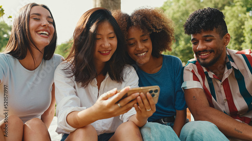 Happy, smiling multiethnic young people at picnic on summer day outdoors. Group of friends talking, using cellphone while relaxing in the park at picnic