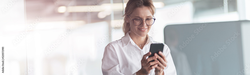 Businesswoman wearing glasses with mobile phone in hand standing in office. Blurred background