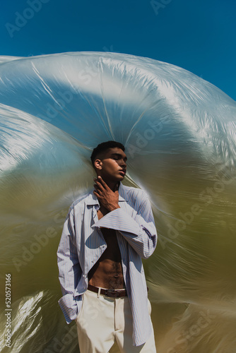 fashionable african american man with closed eyes and hand in pocket touching neck under polyethylene film outdoors