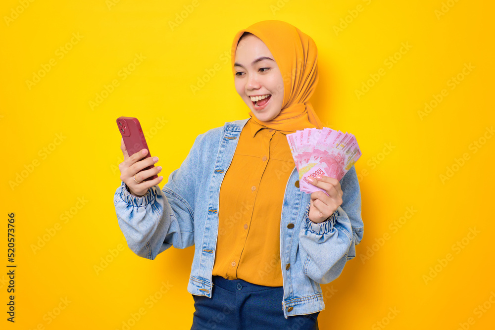 Excited young Asian woman in jeans jacket holding mobile phone and rupiah banknotes, reacting to online win isolated over yellow background Excited young Asian woman in jeans jacket holding mobile phone and rupiah banknotes, reacting to online win isolated over yellow background