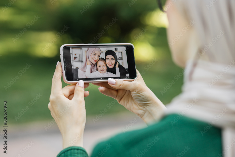 Over the shoulder view of smart phone screen with lovely Muslim multigenerational family, grandmother, mother and little child girl, having a video call with young Arab woman relaxing at park.