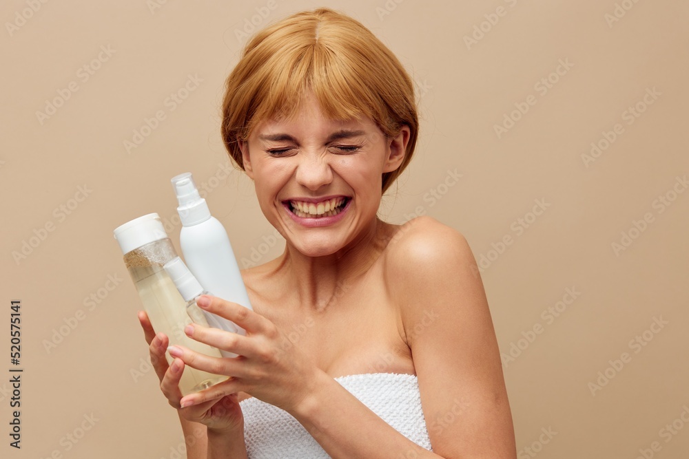 young woman stands on a beige background holding various jars of cosmetics in her hands and smiling radiantly with laughter closes her eyes. photo with empty space for inserting an advertising layout young woman stands on a beige background holding various jars of cosmetics in her hands and smiling radiantly with laughter closes her eyes. photo with empty space for inserting an advertising layout