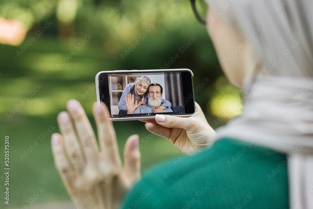 Back view of young muslim woman in hijab, enjoying rest at park, having video call with her parents or grandmother and grandfather, sharing positive emotions Back view of young muslim woman in hijab, enjoying rest at park, having video call with her parents or grandmother and grandfather, sharing positive emotions