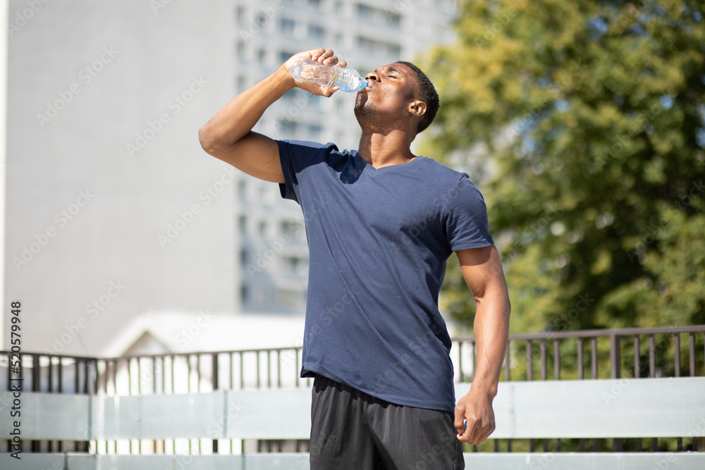 Naklejka premium Thirsty young sportive strong tall handsome African American black skin man drinks water from a bottle, green tree is on background, hot summer day in the city. Horizontal plane. High quality photo.