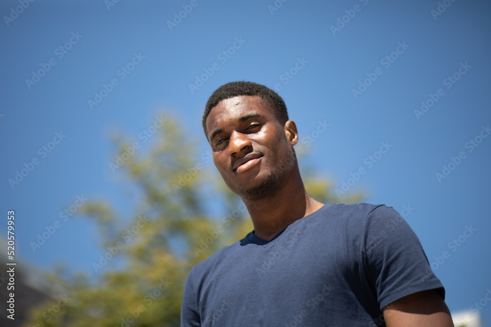 Portrait of young sportive smiling tall handsome African American black skin man with blue sky and green tree on background, hot summer day in urban area. Horizontal plane. High quality photo. Portrait of young sportive smiling tall handsome African American black skin man with blue sky and green tree on background, hot summer day in urban area. Horizontal plane. High quality photo.