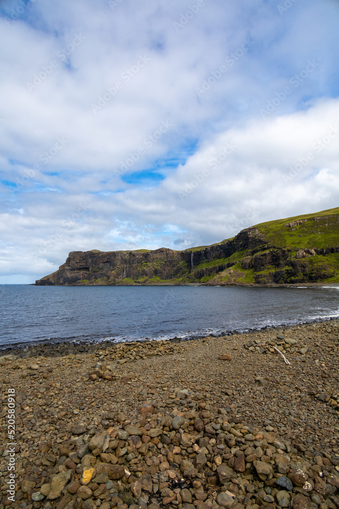 Wide angle views of Talisker Bay, Isle of Skye, Scotland, with its rocky beach, black stones, green fields, and a waterfall at the end of beach. Scattered clouds on blue sky, summer scottish weather.