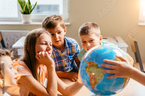 Cheerful curious pupils playing with globe in classroom during geography lesson. Sun glare effect.