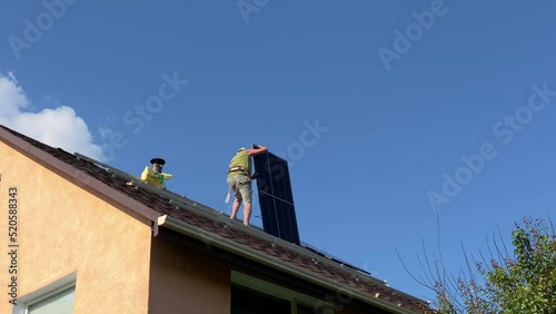 Man carries solar panel for installation on roof of his yellow house secured with rope. Do it yourself DIY PV panels montage on bituminous tiles roof