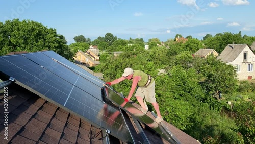 Worker carefully steps on roof to install new solar panels being secured with rope. DIY PV panels setup on bituminous tiles roof