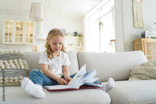 Little european girl with book has leisure at cozy home. Preschool child is learning to read.