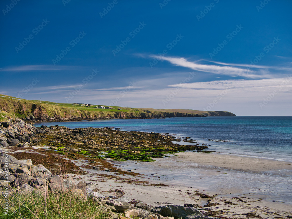 From the beach at Sandwick the picturesque coast of No Ness stretches into the distance. Taken on a sunny day with a blue sky and  high white clouds.