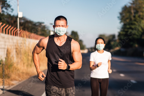 Athletic Asian couple in medical mask jogging on the road together and  looking at camera during covid-19 or coronavirus outbreak. Stay in fit concept.