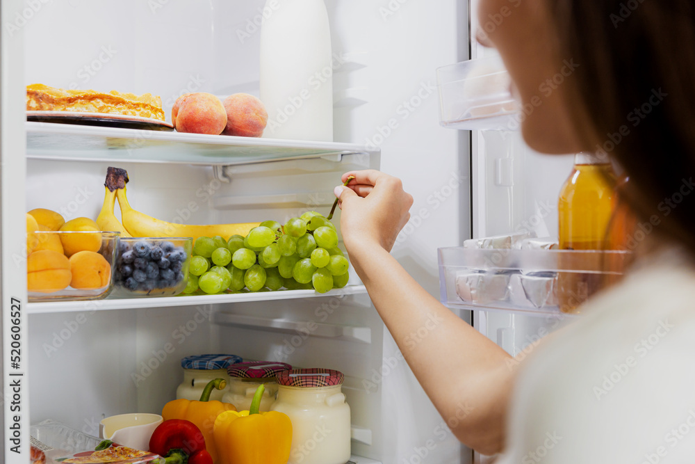 Woman hand taking, grabbing or picks up green bunch of grapes out of open refrigerator shelf or fridge drawer full of fruits, vegetables, banana, peaches, yogurt. Healthy food diet, lifestyle concept Woman hand taking, grabbing or picks up green bunch of grapes out of open refrigerator shelf or fridge drawer full of fruits, vegetables, banana, peaches, yogurt. Healthy food diet, lifestyle concept