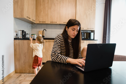 Freelancer woman sits by the table in the home kitchen office, working on laptop. Playful child distracts from work, kid making noise and asking attention from busy mom