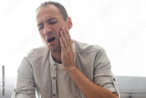 Male patient with severe toothache touching cheek with grimace of pain. Young man with acute dental abscess or inflamed tooth nerve suffering at home. Concept of people having teeth problems.