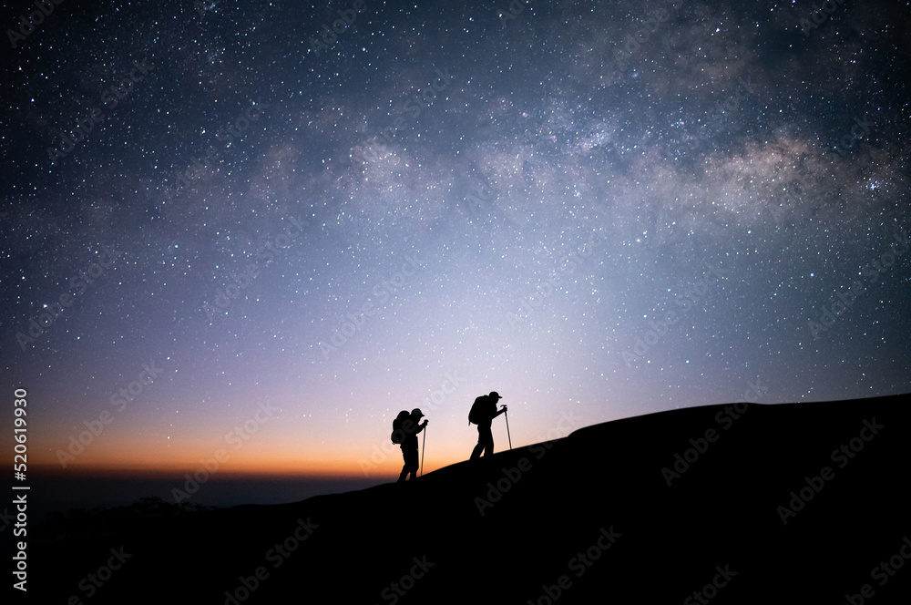 Silhouette of two young traveler and backpacker hiking to the top of the mountain with beautiful view star, milky way over the sky. He enjoyed traveling and was successful when he reached the summit.