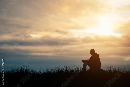 Silhouette of young man sits praying alone at the top of the mountain at sunset with beautiful natural sunlight.