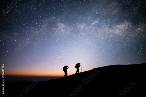 Silhouette of two young traveler and backpacker hiking to the top of the mountain with beautiful view star  milky way over the sky. He enjoyed traveling and was successful when he reached the summit.