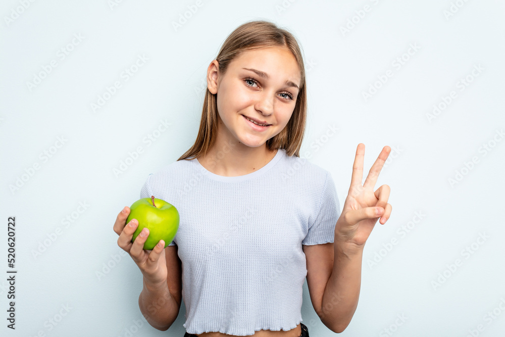 Young caucasian girl holding an apple isolated on blue background joyful and carefree showing a peace symbol with fingers. Young caucasian girl holding an apple isolated on blue background joyful and carefree showing a peace symbol with fingers.