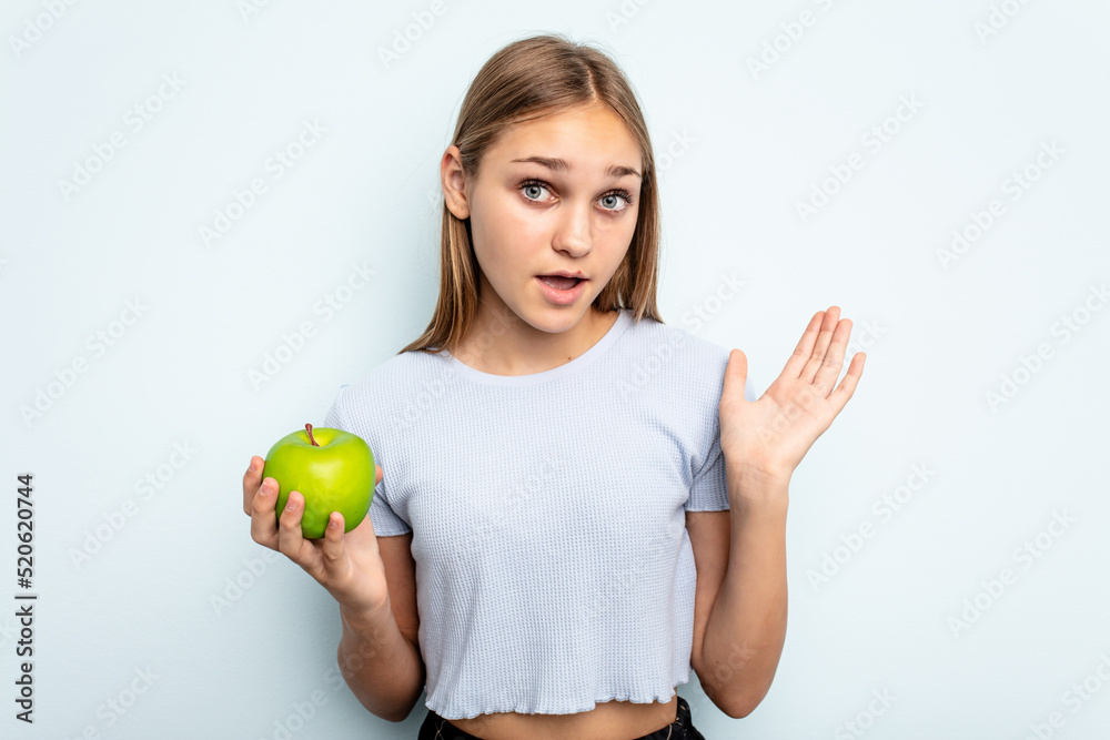 Young caucasian girl holding an apple isolated on blue background surprised and shocked. Young caucasian girl holding an apple isolated on blue background surprised and shocked.