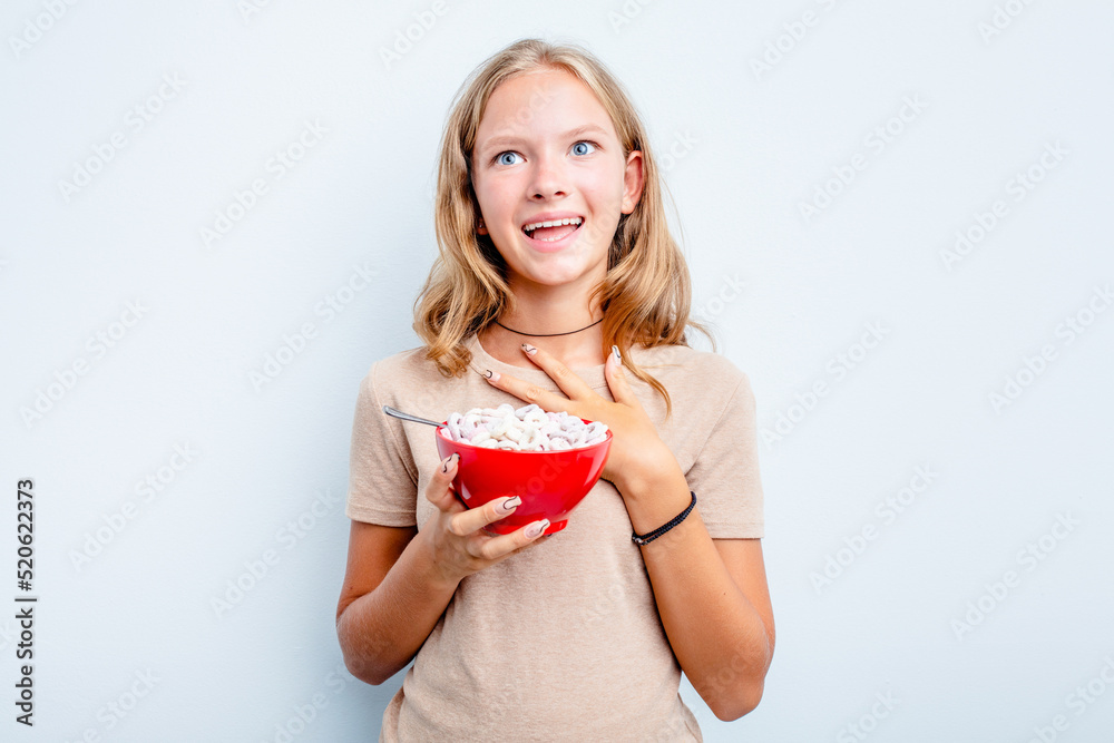 Caucasian teen girl eating cereals isolated on blue background laughs out loudly keeping hand on chest. Caucasian teen girl eating cereals isolated on blue background laughs out loudly keeping hand on chest.