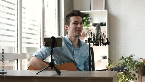 Young male blogger recording video on smartphone web camera, sharing creative hobby. Professional musician playing guitar, singing song, creating tutorial lesson online
