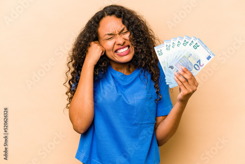 Young hispanic woman holding bank notes isolated on beige background covering ears with hands.