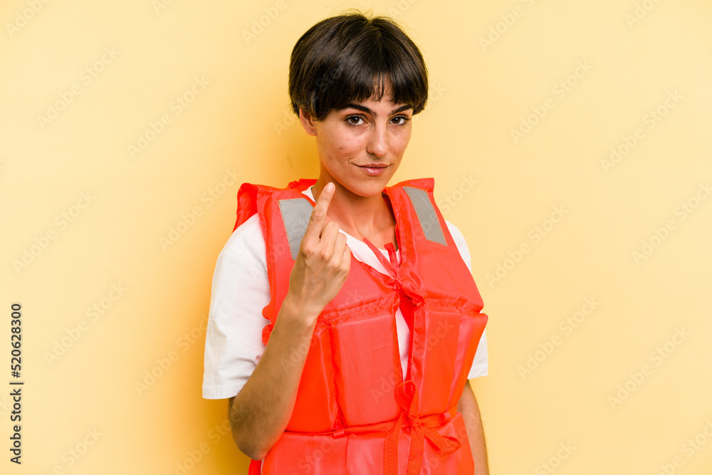 Young caucasian woman wearing life jacket isolated on yellow background pointing with finger at you as if inviting come closer. Young caucasian woman wearing life jacket isolated on yellow background pointing with finger at you as if inviting come closer.