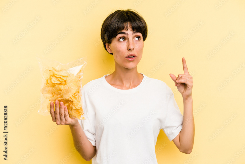Young caucasian man holding crisps isolated on yellow background pointing upside with opened mouth. Young caucasian man holding crisps isolated on yellow background pointing upside with opened mouth.