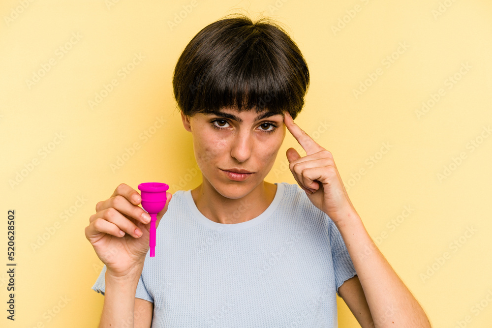 Young caucasian woman holding a menstrual cup isolated on yellow background pointing temple with finger, thinking, focused on a task. Young caucasian woman holding a menstrual cup isolated on yellow background pointing temple with finger, thinking, focused on a task.