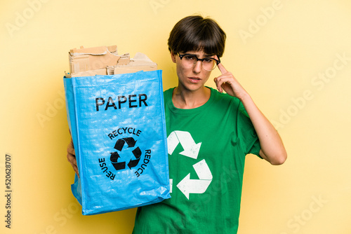 Young woman holding a recycling bag full of paper to recycle isolated on yellow background showing a disappointment gesture with forefinger.