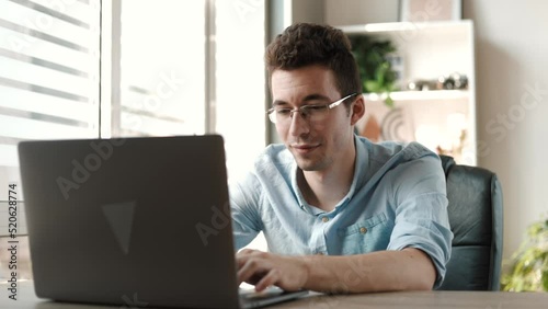 Smiling young businessman working on laptop computer at office. Male professional typing on laptop keyboard at office workplace. Portrait of positive business man looking at laptop screen indoors
