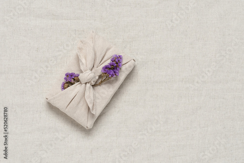 Gift wrapped in fabric with sea lavender, beige textile linen background. Gift in furoshiki style. Zero waste concept. Top view, flat lay, selective focus.