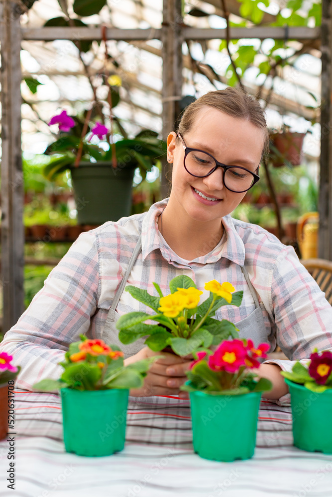 Vertical photo of a young woman in eyeglasses with a few flowers in a greenhouse - she looking at her flowers and smiling.