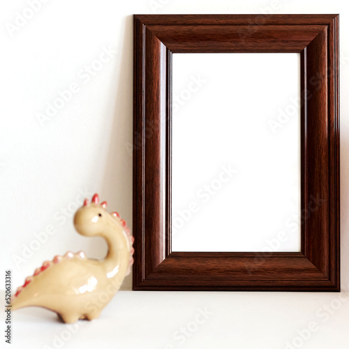 Blank wooden vertically oriented photo frame with funny ceramic dinosaur. Still life, interior, home, style concept background. Mockup, on white background