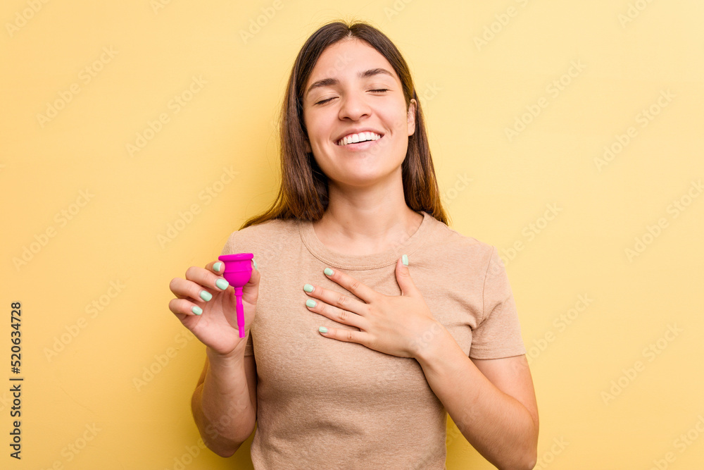 Obraz premium Young caucasian woman holding a menstrual cup isolated on yellow background laughs out loudly keeping hand on chest.