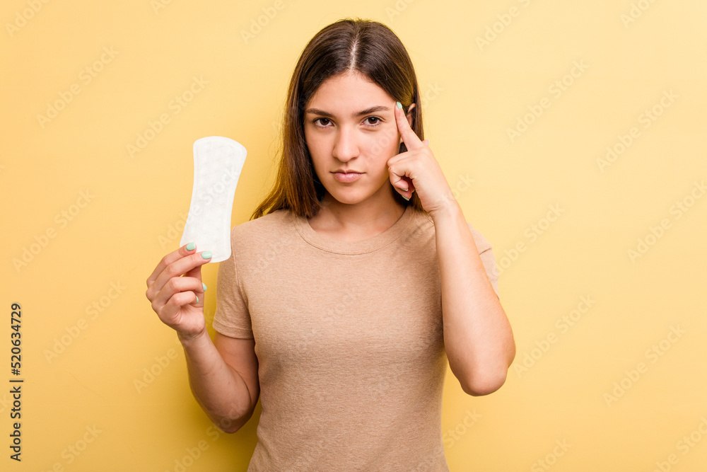 Young caucasian woman holding a sanitary napkin isolated on yellow background pointing temple with finger, thinking, focused on a task. Young caucasian woman holding a sanitary napkin isolated on yellow background pointing temple with finger, thinking, focused on a task.